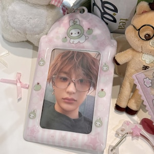 May include: A pink photo frame with a picture of a person wearing glasses. The frame has a cartoon design with stars and a bunny character. Other plush toys and decorative elements are in the background.