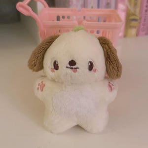 May include: A plush, white dog-like character with brown ears and a green stem. The character has pink blush marks on its cheeks and paws. A small pink shopping cart is in the background.