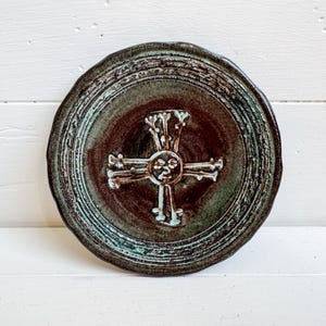 Puede incluir: Un plato redondo de cerámica con un diseño de cruz. El plato presenta un centro marrón oscuro con una cruz blanca en relieve. El borde exterior tiene un tono verde-azulado. El plato tiene un aspecto rústico y hecho a mano.