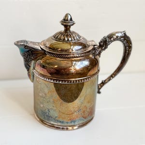 Könnte beinhalten: Antike silberbeschichtete Teekanne mit dekorativem Griff und Deckel. Die Teekanne hat eine angelaufene, schillernde Oberfläche mit blauen und goldenen Farbtönen. Ausguss und Griff weisen aufwendige Details auf.