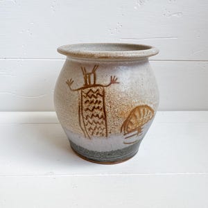 May include: A handcrafted, beige ceramic vase with a wide opening and a rounded body. The vase features a brown, stylized figure and a mushroom design. The pottery has a rustic, earthy aesthetic, perfect for home decor.