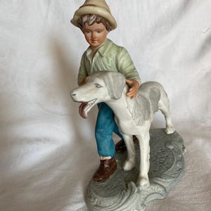 Può includere: Una statuetta in ceramica raffigurante un giovane con una camicia verde, pantaloni blu e un cappello, che tiene un cane. Il cane è bianco con macchie grigie. La statuetta è su una base grigia.
