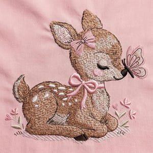 Puede incluir: Imagen bordada de un cervatillo con un lazo rosa en la cabeza y el cuello, y una mariposa en la nariz. El cervatillo es marrón con manchas blancas y está tumbado sobre un fondo rosa. Flores rosas bordadas cerca del cervatillo.