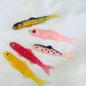 May include: Five colorful, fish-shaped fishing lures. The lures are in shades of yellow, pink, red, and gold, with black and blue accents. Each lure has a detailed eye and fin design, and they are arranged on a white background.
