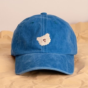 May include: A blue baseball cap featuring a white bear patch. The cap has a curved peak and a button on top. The hat is made from a soft, textured material.
