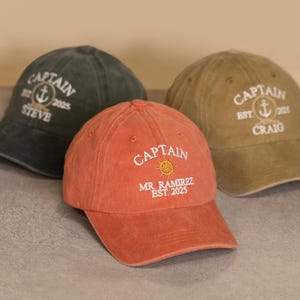 Może przedstawiać: Trzy czapki bejsbolowe w odcieniach zieleni, pomarańczy i beżu. Każda czapka ma wyhaftowane słowo "CAPTAIN" w kolorze białym, motyw marynistyczny i imię. Pomarańczowa czapka wyświetla również "MR. RAMIREZ EST. 2025".