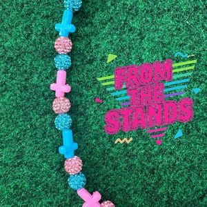 May include: A necklace with pink cross-shaped beads, alternating with sparkly pink and blue round beads. The necklace is on a green background with the text "FROM THE STANDS" in a colorful, retro font.