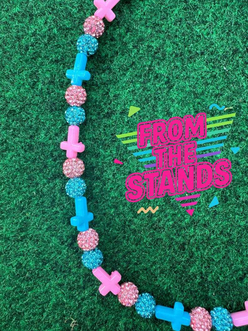 May include: A necklace with pink cross-shaped beads, alternating with sparkly pink and blue round beads. The necklace is on a green background with the text "FROM THE STANDS" in a colorful, retro font.