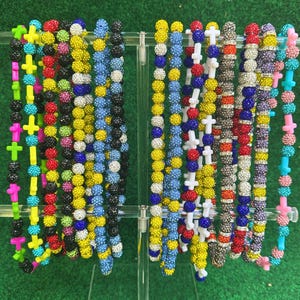 May include: A display of colorful beaded necklaces with cross pendants. The necklaces feature a variety of colors, including yellow, pink, blue, black, and red. The beads are small and round, and the crosses are a contrasting color.