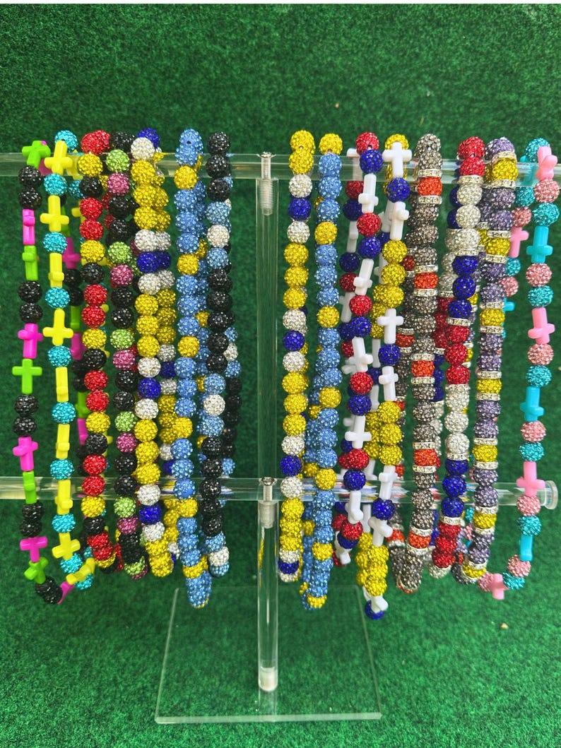 May include: A display of colorful beaded necklaces with cross pendants. The necklaces feature a variety of colors, including yellow, pink, blue, black, and red. The beads are small and round, and the crosses are a contrasting color.