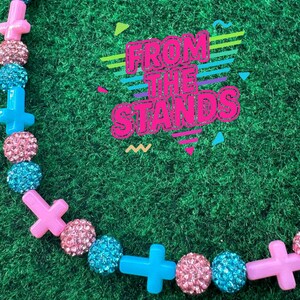 May include: A colorful necklace with pink, blue, and sparkly beads and cross-shaped charms. The necklace is set against a green background. The text "FROM THE STANDS" is displayed in a retro font.