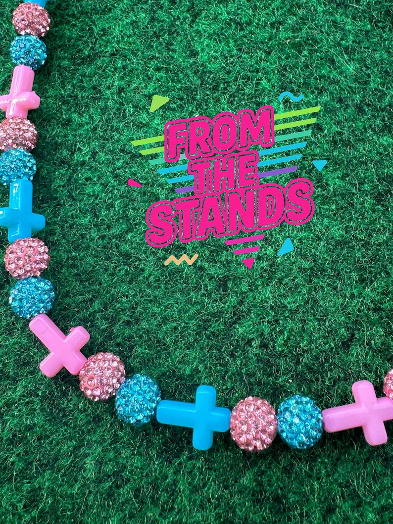 May include: A colorful necklace with pink, blue, and sparkly beads and cross-shaped charms. The necklace is set against a green background. The text "FROM THE STANDS" is displayed in a retro font.
