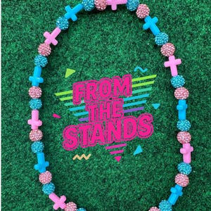 May include: A necklace with alternating pink cross-shaped beads, sparkly pink beads, and sparkly blue beads. The necklace is on a green background. The text "FROM THE STANDS" is in the center of the image.