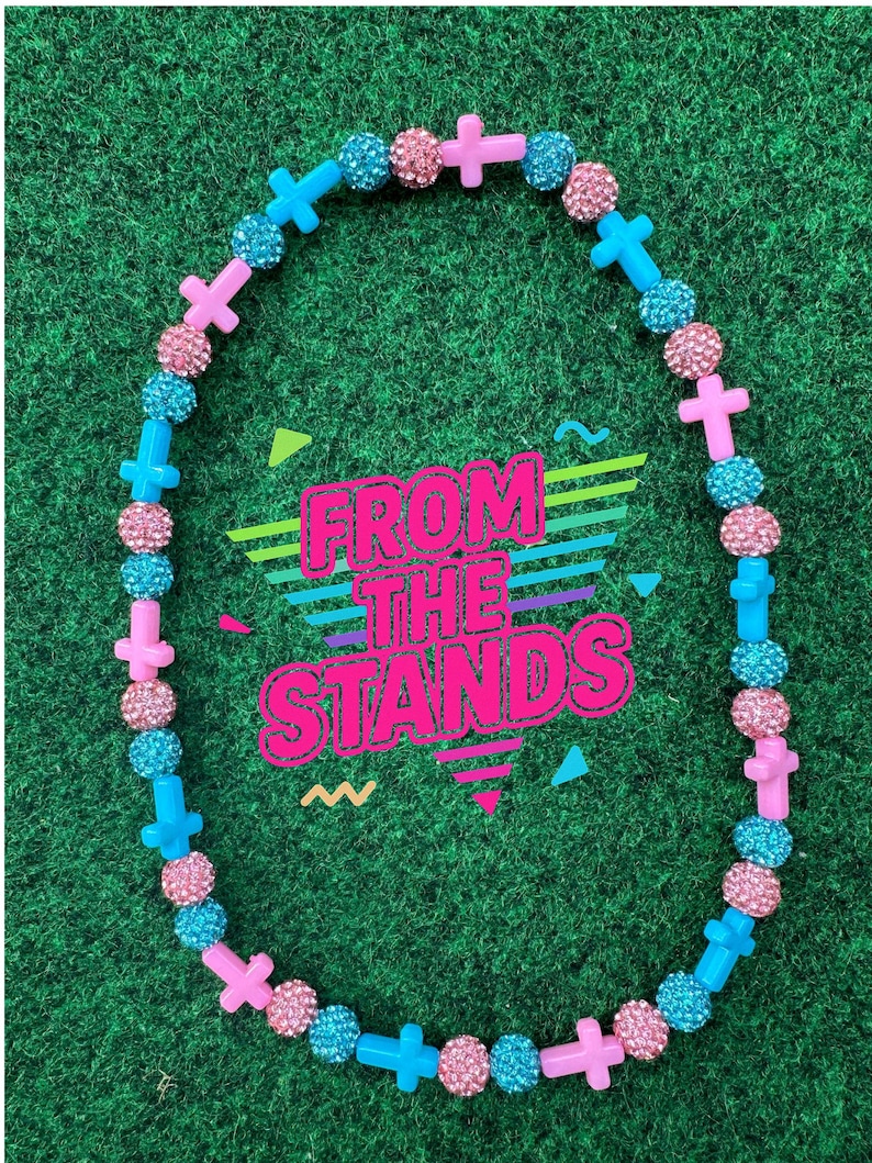 May include: A necklace with alternating pink cross-shaped beads, sparkly pink beads, and sparkly blue beads. The necklace is on a green background. The text "FROM THE STANDS" is in the center of the image.