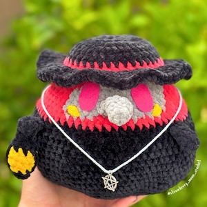 May include: A plush crochet figure of a plague doctor wearing a black wide-brimmed hat with red trim, a gray beak mask, and pink eyes. The figure has a black body, yellow accents on the hands, and a silver pentagram pendant on a white cord.