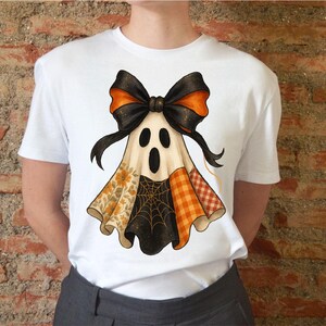 Könnte beinhalten: Weißes T-Shirt mit einem Halloween-Geister-Design. Der Geist hat eine schwarze Schleife mit orangefarbenen Akzenten, einen Patchwork-Körper mit Blumen-, Karo- und Spinnennetzmustern. Das T-Shirt wird von einer Person getragen.