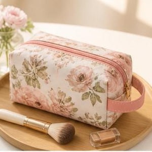 Può includere: Un beauty case con stampa floreale, cerniera e manico rosa è appoggiato su un vassoio di legno. Un pennello per il trucco con manico in legno e un piccolo contenitore trasparente sono accanto alla borsa. La borsa presenta un motivo di rose rosa e foglie verdi.