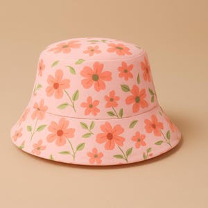 Könnte beinhalten: Ein rosa Fischerhut mit einem sich wiederholenden Muster aus orangefarbenen Blumen und grünen Blättern. Der Hut hat eine abgerundete Krone und eine nach unten geneigte Krempe. Das florale Design bedeckt den gesamten Hut.