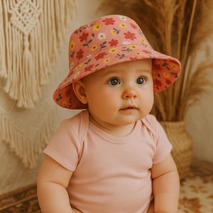 May include: A baby wearing a pink floral print bucket hat and a matching pink onesie. The hat features a repeating pattern of red, yellow, and white flowers. The baby is sitting on a patterned rug.