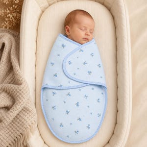 Puede incluir: Un bebé durmiendo envuelto en una manta de arrullo de color azul claro con un pequeño estampado floral en azul oscuro y verde. El arrullo tiene una forma redondeada con un borde de puntos azules y blancos que asegura al bebé.