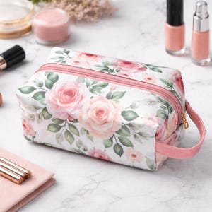May include: A rectangular cosmetic bag featuring a rose floral design in pink and white. It has a pink zip and handle. The background includes makeup items such as nail varnish and jars of cream.