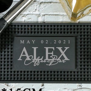 Könnte beinhalten: Eine schwarze Bar-Matte mit strukturierter Oberfläche, die Glaswaren, einen Cocktailshaker und eine Flasche enthält. Die Matte trägt den Text "ALEX Coffee Bar" und das Datum "02. Mai 2021". Die Matte misst 45 x 15 cm.