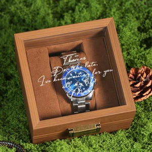 May include: A stainless steel watch with a blue and silver face is displayed inside a wooden box with a brown interior. The watch has a silver band and is set against a brown backdrop. Text on the watch face reads "Thomas, Don't be late. I've been waiting for you all my life, Mary."