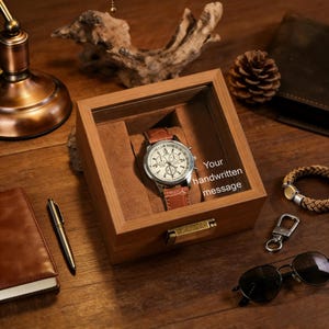 May include: A wooden watch box with a glass top displays a brown leather-banded watch with a white face. The box has a brass clasp and is lined with brown fabric. The text "Your handwritten message" is visible.