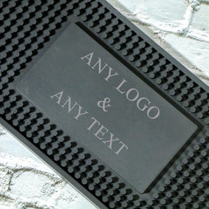 Puede incluir: Una alfombrilla de bar negra con una incrustación rectangular. La incrustación presenta el texto "ANY LOGO & ANY TEXT" en color claro. La alfombrilla tiene una superficie texturizada con pequeñas protuberancias redondeadas.