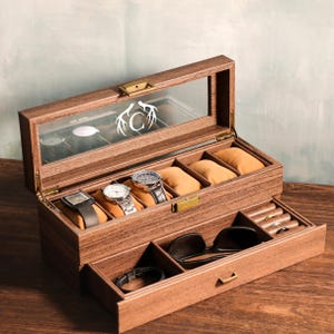 May include: A wooden watch box with a glass lid, showcasing several watches on tan cushions. The box includes a drawer with sunglasses and a bracelet. The top of the box features a white antler design with the letter "C".