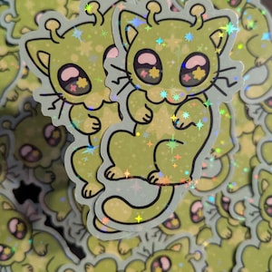 May include: A collection of stickers featuring a cartoon cat-like alien character in shades of green and yellow. The stickers have a holographic, glittery finish with star-shaped accents and large, expressive eyes.