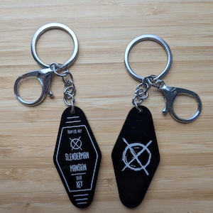 Pode incluir: Dois porta-chaves pretos estilo motel com argolas e fechos de mosquetão em metal prateado. Um apresenta o texto branco "PROXY USE ONLY", um círculo com um X e "SLENDERMAN MANSION DOOR KEY". O outro mostra um círculo branco com um X.