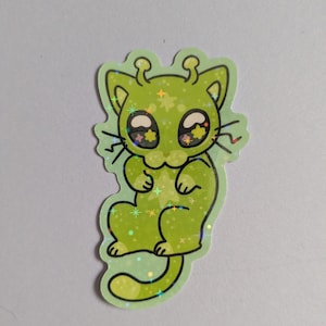 May include: A green cat sticker with a glittery, holographic finish. The cat has large eyes with star-shaped pupils and antennae. The sticker is a cartoon-style illustration of a cat with an alien-like appearance.