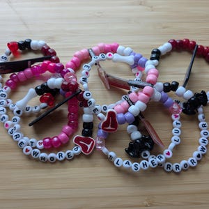 Op de afbeelding: Een verzameling kleurrijke kralenarmbanden met witte, roze, rode, paarse en zwarte plastic kralen. Verschillende armbanden bevatten letterkralen die woorden vormen zoals "SHE GUTS", "SLASHER" en "WHORE", samen met kleine bedels in de vorm van stukjes vlees.