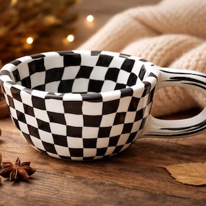 May include: A ceramic coffee mug with a black and white checkered pattern. The mug sits on a wooden surface with a lit candle, cinnamon sticks, and sugar cubes. A cozy, cream-colored blanket is in the background.
