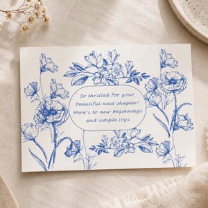 May include: A rectangular white greeting card featuring blue ink botanical illustrations of flowers and leaves. Centered in an oval frame is the text: So thrilled for your beautiful next chapter! Here's to new beginnings and simple joys.