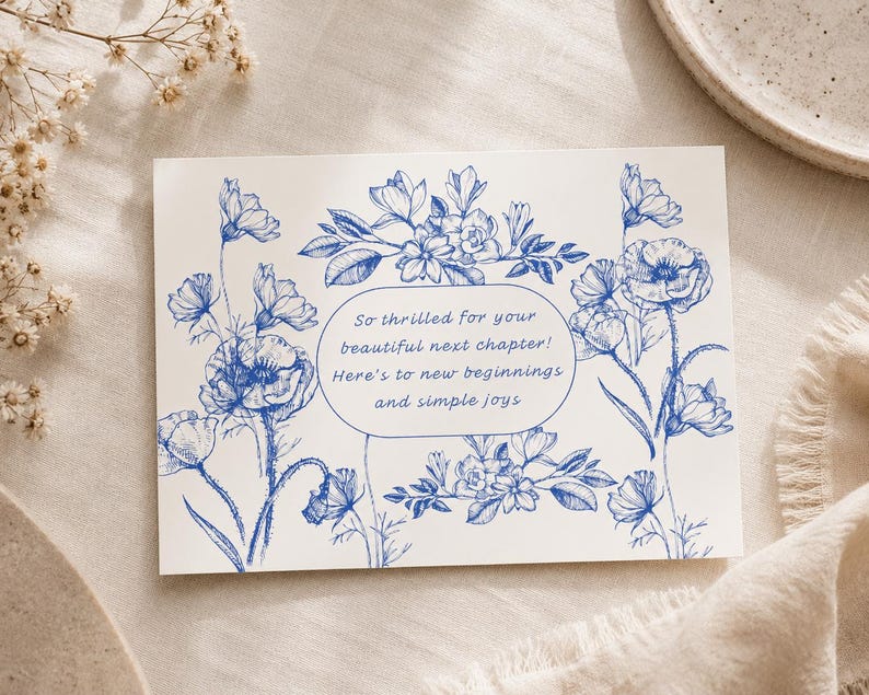 May include: A rectangular white greeting card featuring blue ink botanical illustrations of flowers and leaves. Centered in an oval frame is the text: So thrilled for your beautiful next chapter! Here's to new beginnings and simple joys.