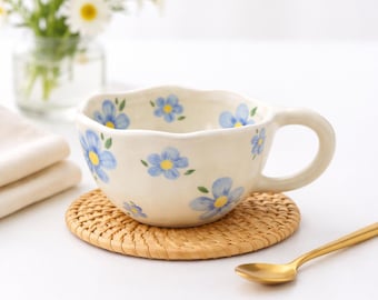 May include: A cream-colored ceramic teacup with blue floral designs and a wavy rim sits on a round, woven coaster. A gold spoon rests nearby. A vase of daisies is in the background.