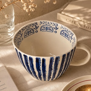May include: A ceramic mug with a cream-colored base featuring hand-painted dark blue vertical stripes on the exterior and a decorative blue pattern along the inner rim. The mug has a curved handle and a wide, rounded bowl shape for holding beverages.