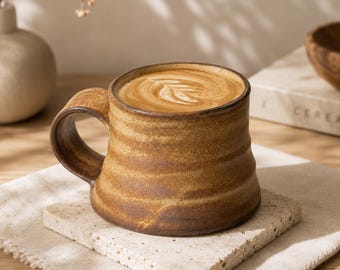 May include: A ceramic mug with a wide, tapered base and a thick handle, featuring a textured, mottled brown and tan glaze. The mug is filled with a coffee beverage topped with a white leaf-shaped latte art design on the surface.