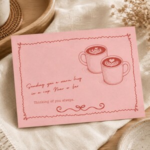May include: A rectangular pink greeting card featuring a red line-art illustration of two coffee mugs with latte art. The text reads: Sending you a warm hug in a cup. Near or far. Thinking of you always. The card has a decorative scribbled border.