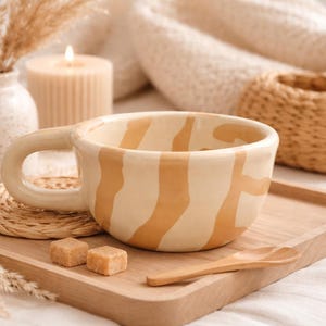 May include: A ceramic mug with a wide, rounded handle and a cream-colored base featuring irregular, vertical tan stripes. The mug is positioned on a light-colored wooden tray alongside a small wooden spoon and two brown sugar cubes.