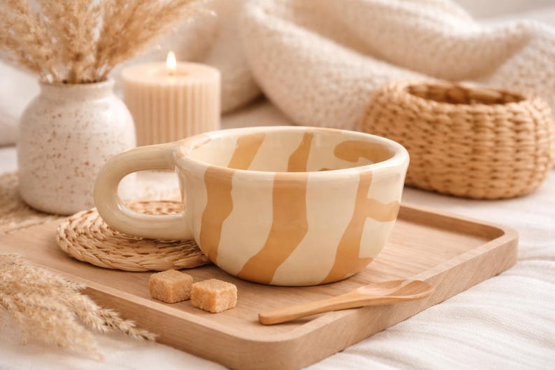 May include: A ceramic mug with a wide, rounded handle and a cream-colored base featuring irregular, vertical tan stripes. The mug is positioned on a light-colored wooden tray alongside a small wooden spoon and two brown sugar cubes.