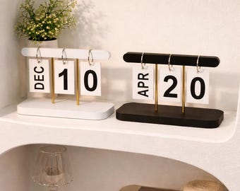 May include: Two wooden perpetual calendars, one white and one black, each displaying a month and date. The white calendar shows "DEC 10", while the black calendar shows "APR 20". Both have gold-colored metal supports and are on a white shelf.
