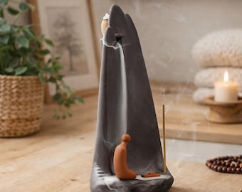 May include: A gray ceramic incense burner with a mountain-like shape. A small terracotta figure is kneeling in front of the mountain, with incense smoke flowing down. A wooden incense stick is placed on the side.