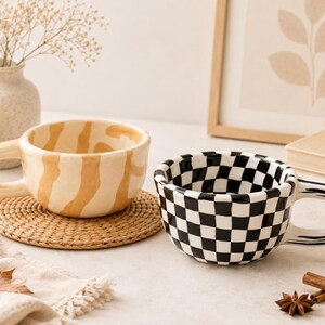 May include: Two ceramic mugs with rounded handles. One mug features a beige and cream wavy striped pattern, while the other displays a black and white checkered pattern. Both mugs have a thick, rounded rim and a smooth, glossy finish.