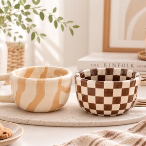 May include: Two ceramic mugs with rounded handles. One mug features a cream base with irregular tan wavy stripes. The second mug displays a brown and white checkered pattern. Both mugs have a glossy finish and a wide, low-profile bowl shape.