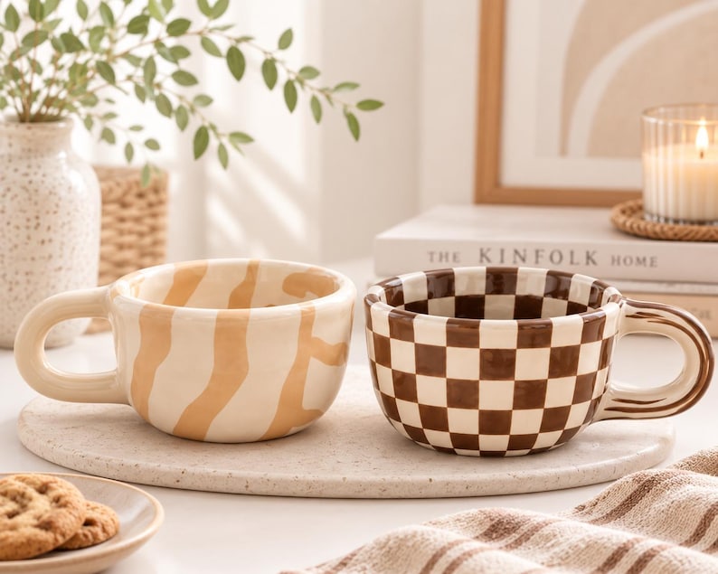 May include: Two ceramic mugs with rounded handles. One mug features a cream base with irregular tan wavy stripes. The second mug displays a brown and white checkered pattern. Both mugs have a glossy finish and a wide, low-profile bowl shape.