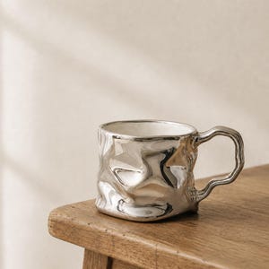 May include: A metallic silver mug with a highly reflective, irregular, and rippled surface texture. The mug features a matching silver handle with a wavy, organic shape and a smooth white interior finish, creating a distinct contrast.