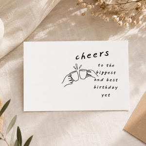 May include: A white rectangular greeting card featuring a black line drawing of two hands clinking mugs together. The text on the card reads: cheers to the biggest and best birthday yet. The design is centered on the front of the card.